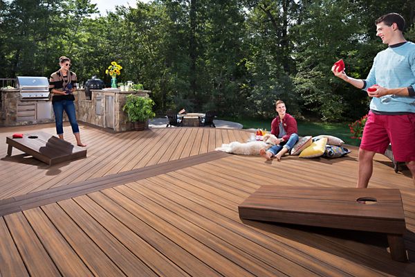 A group of adults enjoys a game of cornhole on a large, modern wooden deck surrounded by greenery. The setting features an outdoor kitchen area with a grill and stone counter, and a dog lounges beside two people seated on the deck. The atmosphere is casual and social, with bright daylight and a relaxed mood.