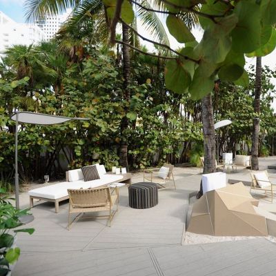 Contemporary outdoor lounge seating is arranged on a light wooden deck surrounded by lush tropical greenery and tall palm trees. White sofas, woven chairs, and low tables create a relaxed social area in a resort-like setting. Geometric beige structures and striped ottomans add visual interest and modern design elements. The scene suggests a tranquil, stylish space for relaxation or events in a warm coastal environment.