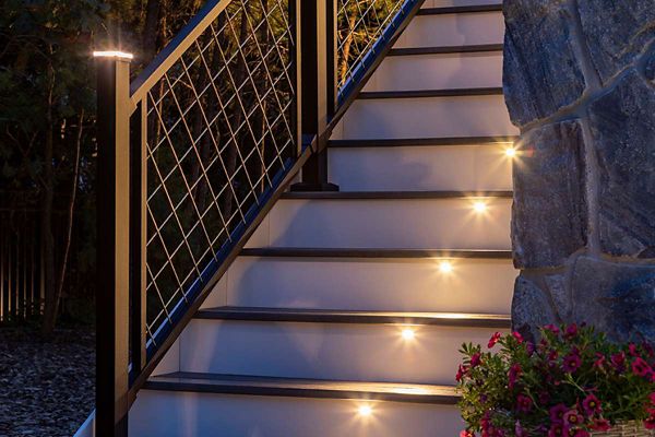 A contemporary outdoor staircase is illuminated by built-in step lights, creating a welcoming and safe ascent. The stairs feature a sleek metal railing with a wire mesh design, set against a stone wall and surrounded by greenery. Warm white lights are embedded in each step, enhancing visibility and ambiance. A planter with pink flowers adds a touch of color to the scene.