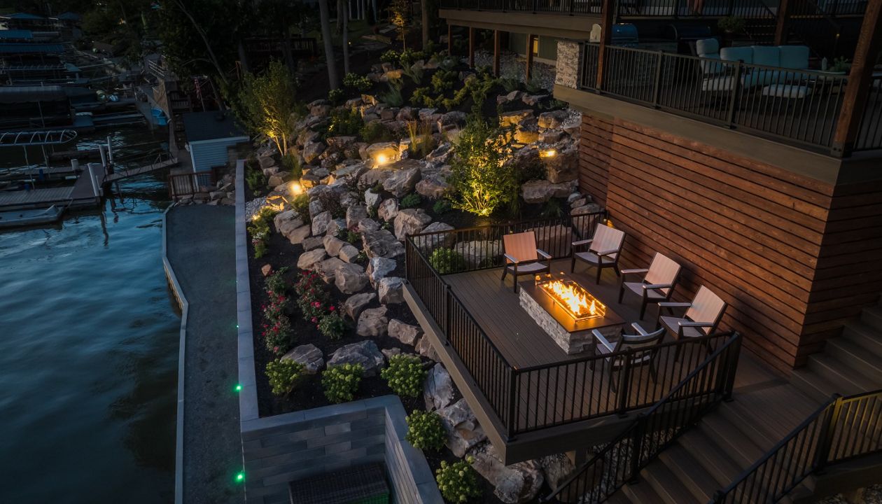 Lower-level composite deck featuring a built-in fire pit seating area surrounded by natural boulder landscaping and shoreline views.