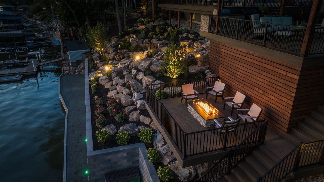 Lower-level composite deck featuring a built-in fire pit seating area surrounded by natural boulder landscaping and shoreline views.