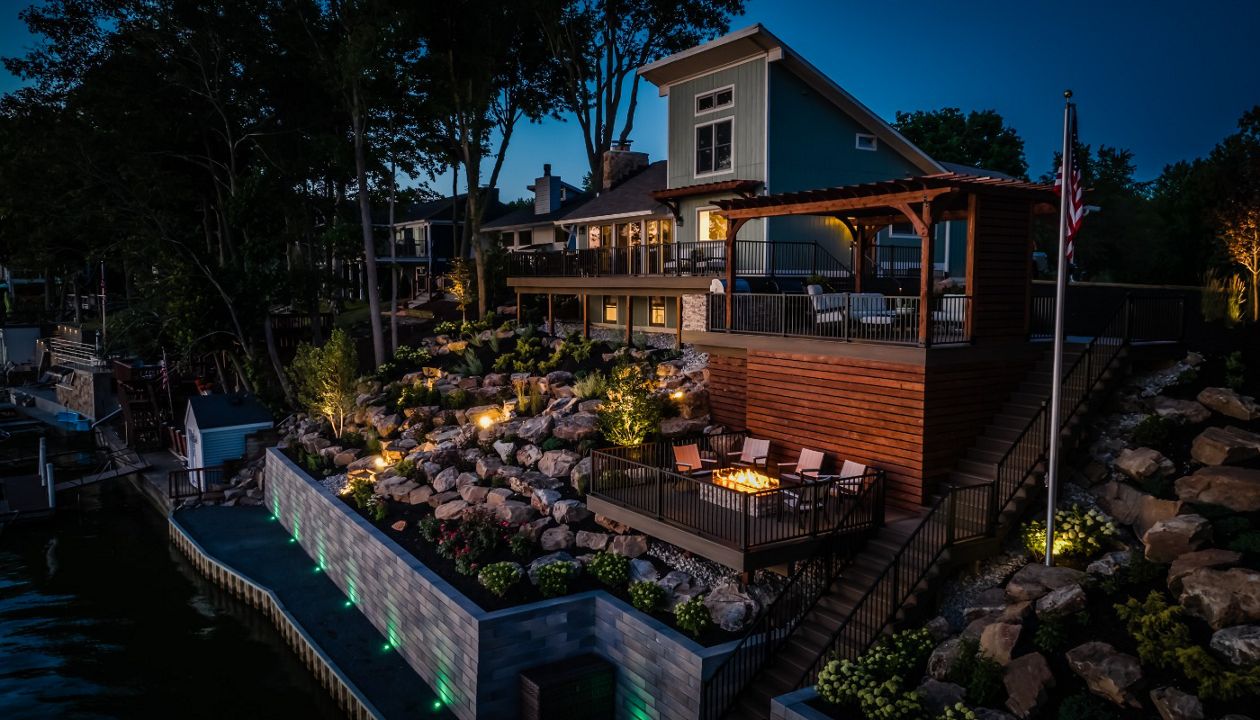 Lakeside composite deck with custom pergola, integrated fire pit lounge, and tiered landscape lighting overlooking the water.