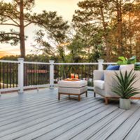 A contemporary outdoor deck features wicker lounge chairs and a small table, set against a scenic lakeside view at sunset. The deck is accented with decorative pillows, a potted plant, and candles, creating a cozy and inviting atmosphere. The railing provides a clear view of the tranquil water and surrounding trees, while the warm sunset light enhances the peaceful mood.