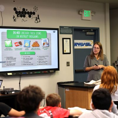 A teacher stands at the front of a classroom giving a presentation to a group of students. A large screen displays a slide about items not to include in a recycling or waste program, featuring illustrated examples of various materials. The classroom setting includes lab tables, a door with a sign, and educational wall graphics. Students of different ages sit facing the screen, listening to the lesson. The overall scene conveys an educational, informative atmosphere focused on environmental awareness.