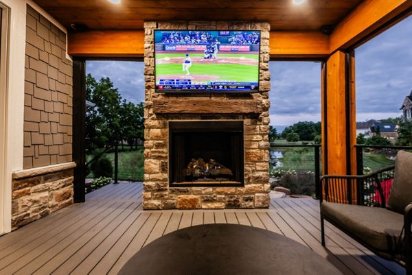 Covered deck lounge featuring a stone fireplace, mounted TV, and warm wood ceiling accents.