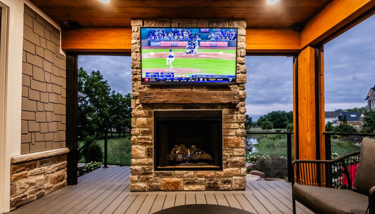 Covered deck lounge featuring a stone fireplace, mounted TV, and warm wood ceiling accents.