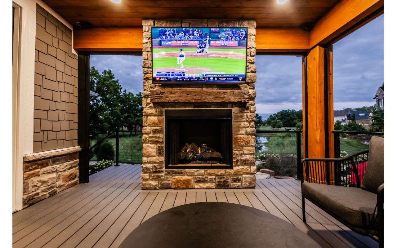 Covered deck lounge featuring a stone fireplace, mounted TV, and warm wood ceiling accents.