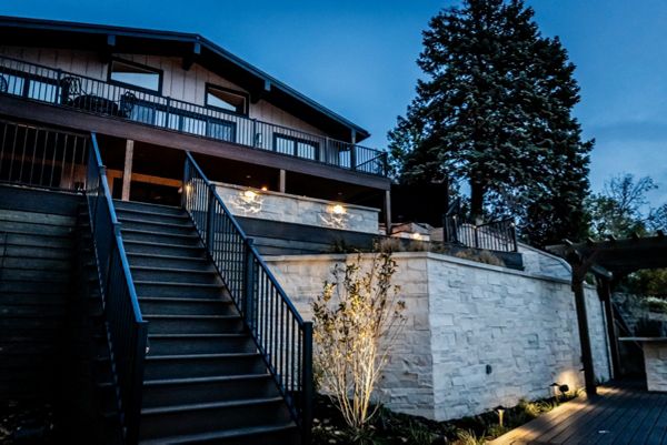 Wide composite deck staircase with aluminum railing connecting the upper outdoor living space to the lower patio, highlighted by integrated landscape and accent lighting.