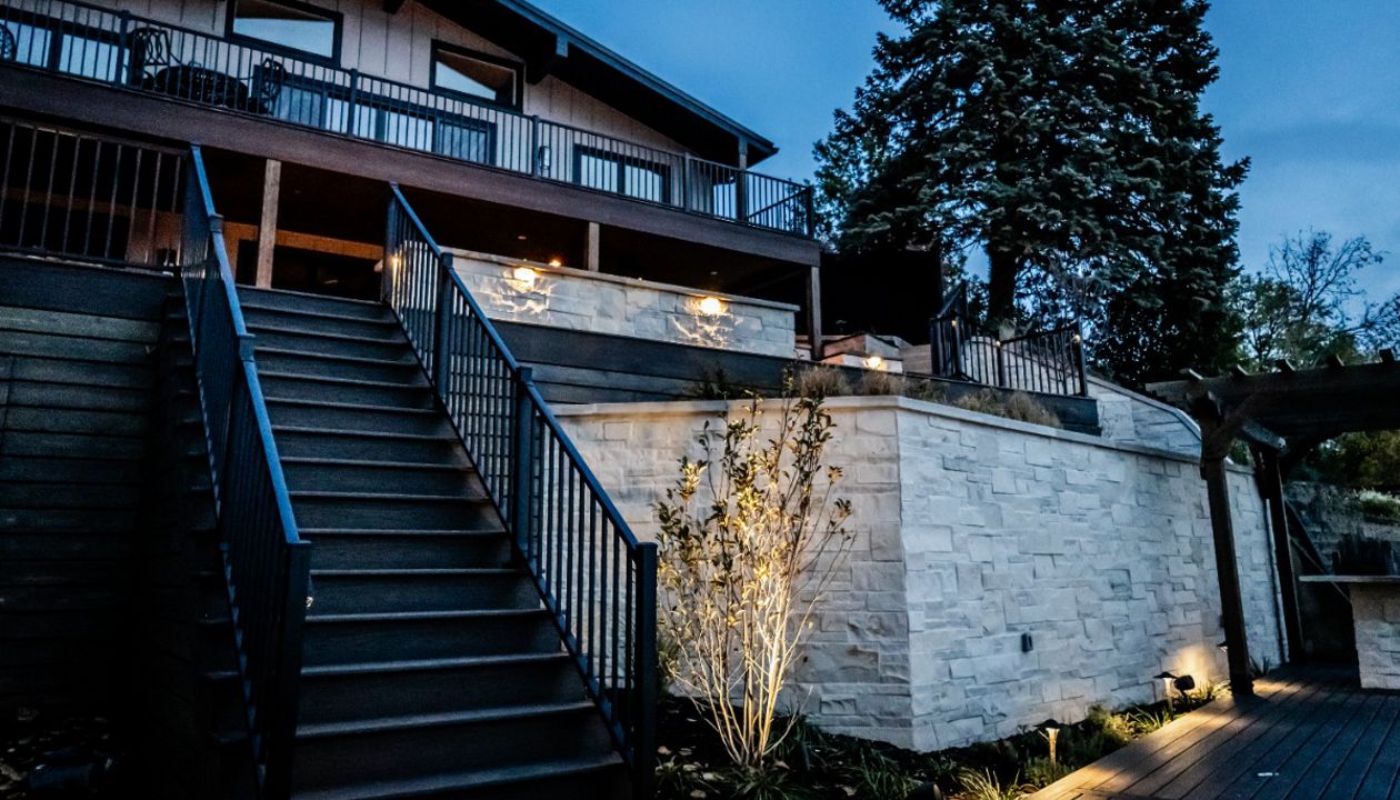 Wide composite deck staircase with aluminum railing connecting the upper outdoor living space to the lower patio, highlighted by integrated landscape and accent lighting.