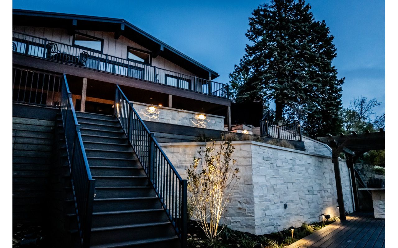 Wide composite deck staircase with aluminum railing connecting the upper outdoor living space to the lower patio, highlighted by integrated landscape and accent lighting.