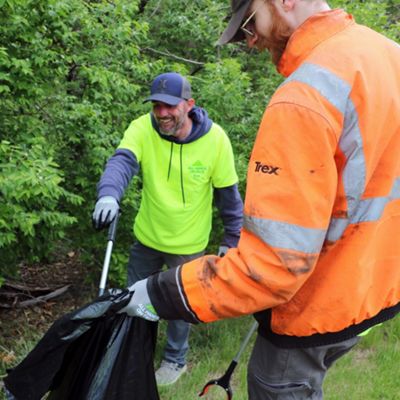 Two adults are picking up trash along a grassy area beside dense green bushes. One person in a bright yellow shirt uses a litter grabber to place waste into a large black garbage bag held by another person in an orange safety jacket. Both wear work gloves and casual outdoor clothing suitable for manual cleanup. The scene suggests a community or environmental volunteer effort focused on keeping nature areas clean.