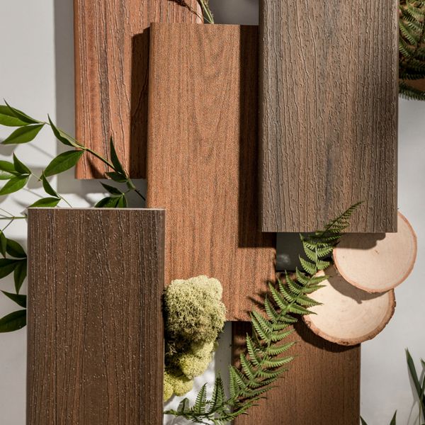 Several rectangular ceramic tiles with a wood grain finish are arranged on a neutral background. The tiles display various shades of brown, mimicking natural wood textures. Green leaves, ferns, and moss are artfully placed around the tiles, along with two round wood slices, creating an organic and earthy setting. The overall mood is natural and rustic, with a focus on eco-friendly design elements.