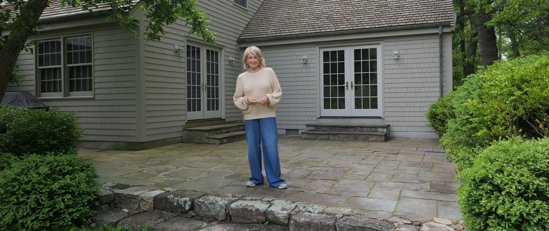 A woman stands on a large stone patio in front of a gray wooden house. The home features multiple windows, a gabled roof, and double glass doors leading to the patio. Lush green shrubs and trees surround the yard, creating a peaceful garden setting. The scene feels calm and relaxed, with soft natural daylight illuminating the outdoor space.