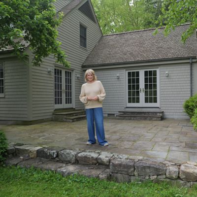 A woman stands on a large stone patio in front of a gray wooden house. The home features multiple windows, a gabled roof, and double glass doors leading to the patio. Lush green shrubs and trees surround the yard, creating a peaceful garden setting. The scene feels calm and relaxed, with soft natural daylight illuminating the outdoor space.