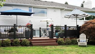 A spacious backyard patio features multiple large umbrellas providing shade over a seating and dining area. The deck is elevated with dark railings and brown steps, surrounded by neatly trimmed bushes and green grass. White and blue striped umbrellas, potted flowers, and an Adirondack chair add a relaxed, summery vibe to the scene.
