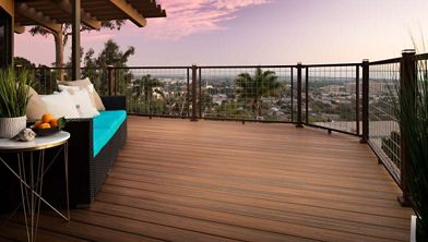 A spacious wooden deck overlooks a cityscape under a vibrant pink and purple sunset sky. The deck features a contemporary outdoor sofa with turquoise cushions and a small side table holding fruit. Glass and metal railings frame the view, while potted plants add a touch of greenery to the relaxing outdoor setting.
