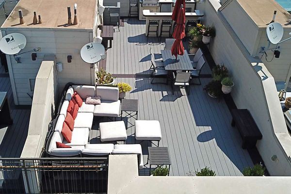 A spacious rooftop patio features a sectional sofa with white cushions and red accent pillows, accompanied by matching ottomans and a small table. The area includes a dining table with red umbrellas, potted plants, and satellite dishes mounted on the surrounding walls. The setting is bright and airy, with a contemporary outdoor style and neutral gray decking.