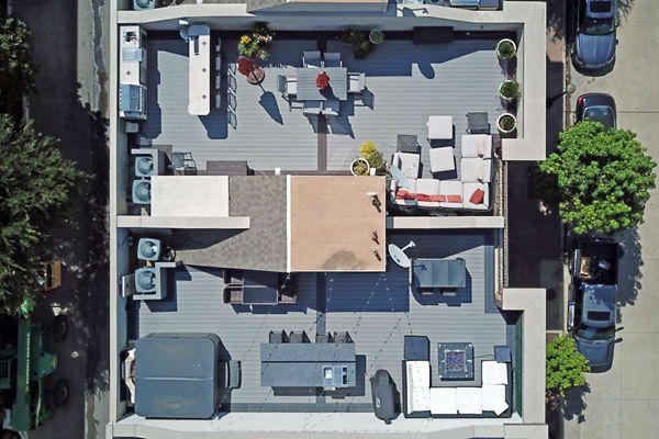 Aerial view of a contemporary rooftop patio featuring multiple seating zones, including lounge chairs, sofas, and dining tables. The setting includes potted plants, outdoor furniture, and a barbecue grill, all arranged on a spacious deck. The area is surrounded by urban buildings and parked cars, with a mix of shaded and sunny spots.