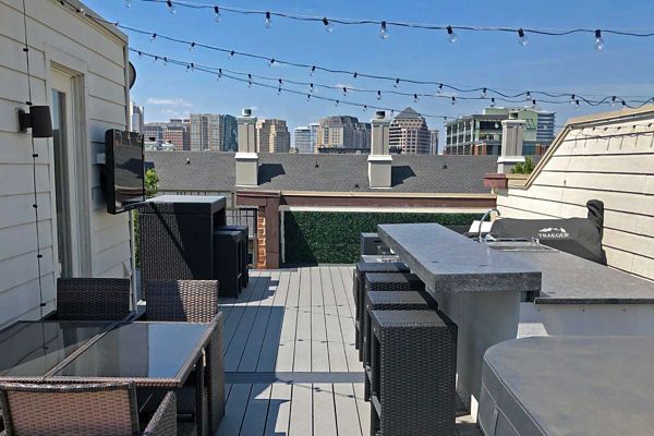 A stylish rooftop patio features wicker furniture, a glass dining table, and a stone bar counter. String lights are suspended overhead, creating a welcoming atmosphere for gatherings. The setting overlooks a city skyline under a clear blue sky, with a mounted television and grill visible.
