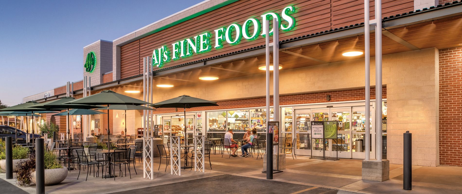 The image shows the exterior of AJ's Fine Foods, a grocery store with a modern facade. Outdoor seating with tables and umbrellas is visible in front of the entrance, suggesting a welcoming atmosphere for customers. The store's name is prominently displayed in green illuminated letters above the entrance. The setting appears to be early evening, with clear skies and warm lighting. No visible numbers or additional text are present.