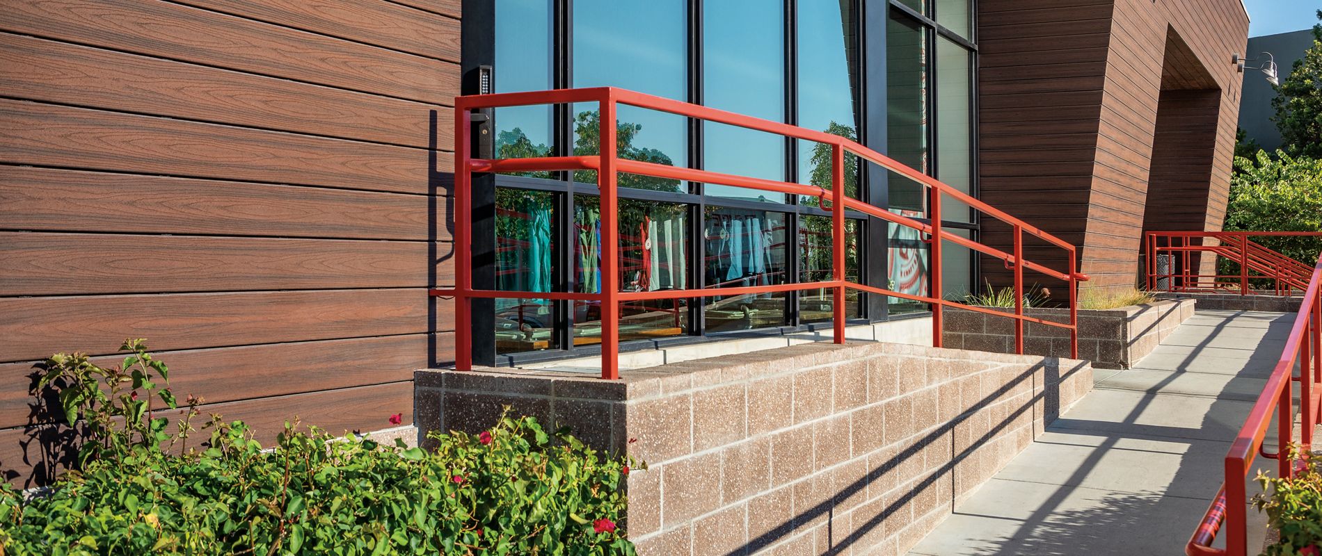 A contemporary commercial building exterior features large reflective windows and wood-textured siding. The entrance area includes a concrete ramp with bright red metal handrails, providing accessibility. Sunlight casts sharp shadows, highlighting the clean architectural lines and landscaped greenery along the walkway. No visible text or numbers are present in the image.