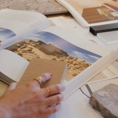 A person with light skin and jewelry on their fingers examines a landscape photo book while holding a textured material sample. The setting appears to be a design studio or workspace, with stone samples and open books spread across a table. The mood is creative and focused, with neutral and earthy tones dominating the scene. No visible text or numbers are present in the image.