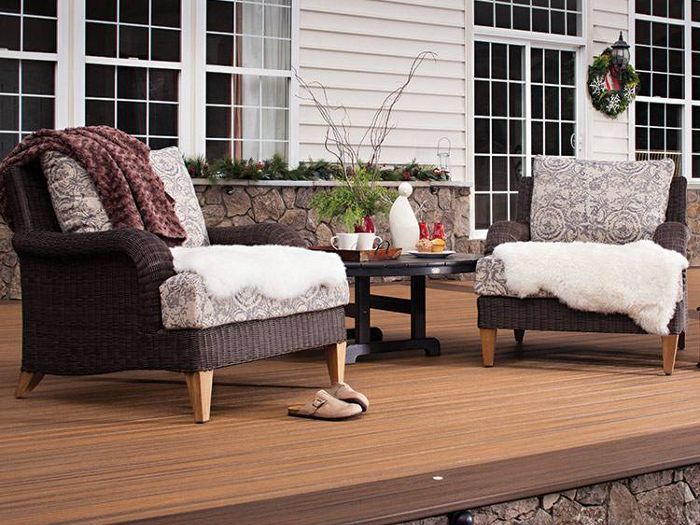 Two cushioned armchairs with patterned upholstery and soft throws are arranged on a wooden patio deck. A round table between the chairs holds a teapot, cups, and a decorative vase, creating a welcoming outdoor setting. The background features a house with large windows, festive wreath, and greenery, suggesting a comfortable and inviting atmosphere.