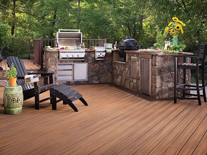 A modern outdoor kitchen features a built-in stainless steel grill and appliances set in a stone counter. The scene is set on a spacious wooden deck surrounded by lush green trees, with Adirondack chairs and a table with sunflowers adding a relaxed, inviting atmosphere. The area is well-organized for outdoor cooking and entertaining, with visible utensils, drinks, and fresh produce.