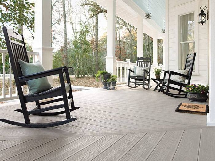 A spacious front porch features several black wooden rocking chairs, a small side table, and potted plants. The porch is attached to a white house with columns and overlooks a wooded yard. The setting is calm and inviting, with soft natural light and a neutral color palette. A doormat is visible near the entrance, adding a welcoming touch.