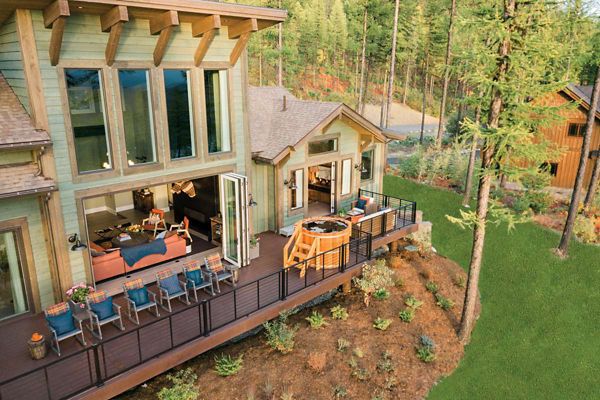 A contemporary house features large windows and an expansive deck overlooking a forested landscape. The outdoor area includes multiple seating arrangements and a round hot tub, perfect for relaxation and gatherings. The home is surrounded by lush greenery and tall trees, creating a tranquil, nature-inspired setting.