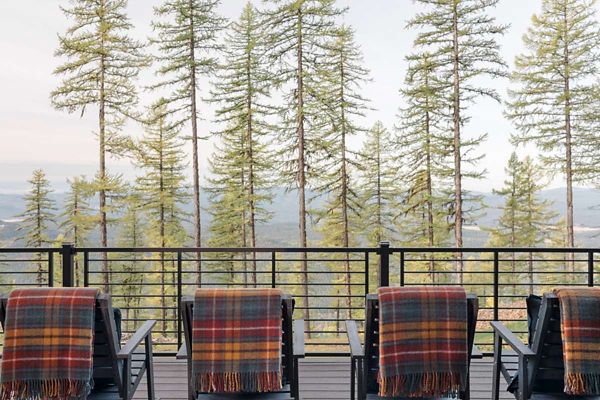 Four outdoor chairs are arranged on a deck, each draped with a colorful plaid blanket. The deck features a metal railing and faces a serene view of tall pine trees and distant mountains. The setting suggests a peaceful, cozy atmosphere ideal for relaxation. The color palette includes earthy tones and soft pastels, enhancing the tranquil mood. No visible text or numbers are present in the image.