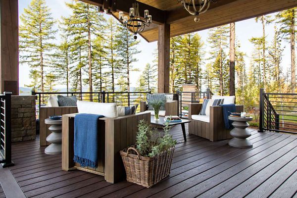 A contemporary outdoor patio features four cushioned armchairs arranged around a small table, set against a scenic forest backdrop. The space is decorated with neutral and blue textiles, a woven basket with greenery, and stylish side tables. Warm wood tones and soft natural light create a relaxing, inviting atmosphere. No visible text or numbers are present in the image.