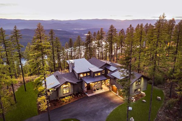 A contemporary house is nestled among tall pine trees on a mountain slope at sunset. The home features multiple rooflines, large windows, and warm exterior lighting, creating a cozy and inviting atmosphere. The setting offers expansive views of distant mountains and valleys, with a winding driveway leading to the entrance. The overall mood is tranquil and secluded, emphasizing natural beauty and modern architecture.