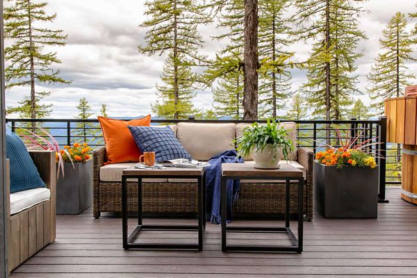 A comfortable outdoor patio features a wicker sofa with colorful cushions and a throw blanket, set against a backdrop of tall pine trees. Two modern black tables hold a mug, book, and a potted plant, while large planters with vibrant flowers add a touch of color. The setting is tranquil and inviting, with a natural palette and a scenic forest landscape visible beyond the railing.