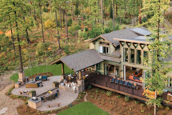 A contemporary multi-level house is nestled in a forested landscape, featuring large windows and a spacious outdoor patio. The patio includes multiple seating areas, a fire pit, and dining furniture, all surrounded by natural greenery and tall trees. The setting suggests a tranquil, rustic retreat with modern amenities and earthy tones.