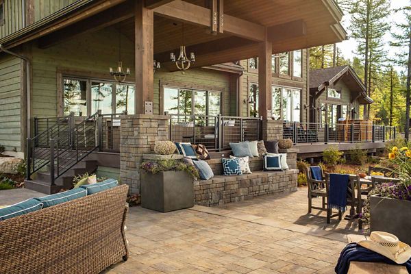 A large, modern patio features comfortable wicker and cushioned seating arranged around a stone fire pit. The setting is a wooded residential area with a contemporary house in the background, large windows, and a covered deck. Decorative pillows, potted plants, and outdoor dining furniture create a welcoming, relaxed atmosphere. Warm earth tones and natural materials dominate the palette, evoking a rustic yet elegant style.