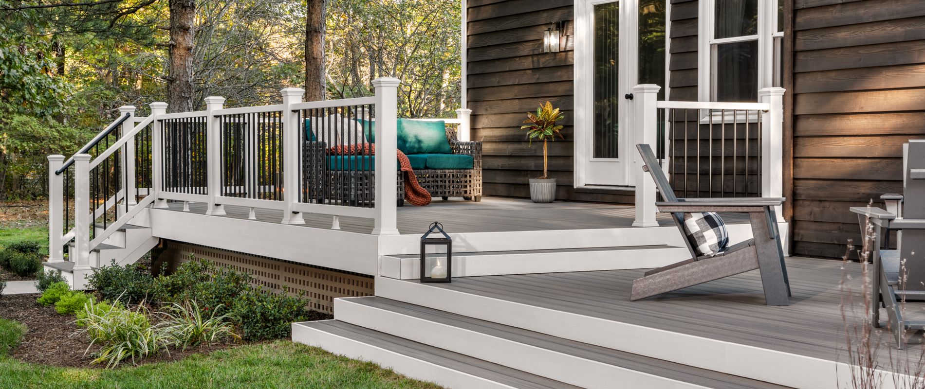A spacious, elevated backyard deck is attached to a dark wood house, featuring white railings and steps. The setting includes a cushioned bench with a person relaxing, and a single lounge chair with a plaid pillow. Surrounded by greenery and trees, the scene evokes a peaceful, contemporary outdoor living space.