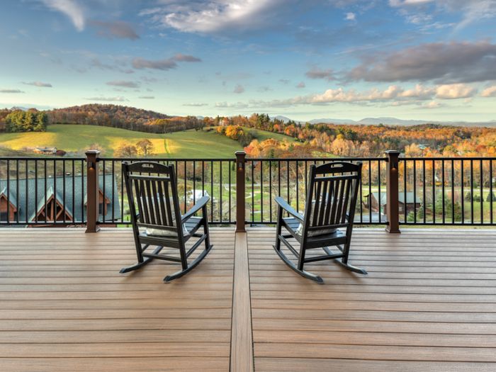 Two empty rocking chairs are positioned on a spacious wooden deck overlooking a picturesque rural landscape. The view features rolling green hills, scattered trees, and a few distant houses under a partly cloudy sky. The scene is bathed in warm, late afternoon light, creating a tranquil and inviting atmosphere. The deck is bordered by a dark railing, framing the expansive countryside.