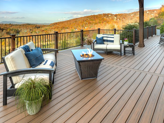 A spacious outdoor deck features cushioned seating and a central fire table, overlooking a scenic mountain landscape. The setting is tranquil, with warm wood-toned flooring and black railings. Neutral and blue accent pillows add a touch of color, while a potted plant brings greenery to the space. The overall mood is inviting and relaxed, ideal for gatherings or quiet moments.