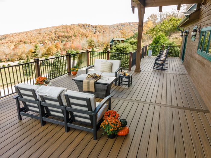 A large wooden deck features comfortable cushioned seating arranged around a central table, set against a scenic backdrop of rolling hills with autumn foliage. Decorative pumpkins and potted orange flowers add a seasonal touch to the space. The deck is bordered by a black railing and extends alongside a house, with additional seating visible in the distance. The setting suggests a peaceful, inviting atmosphere ideal for relaxation or gatherings.