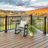 A single black rocking chair with a cushion sits on a wooden deck, accompanied by a potted plant. The deck features black railings and overlooks a scenic valley filled with colorful autumn foliage. The sky is partly cloudy, casting warm light over the tranquil outdoor setting.