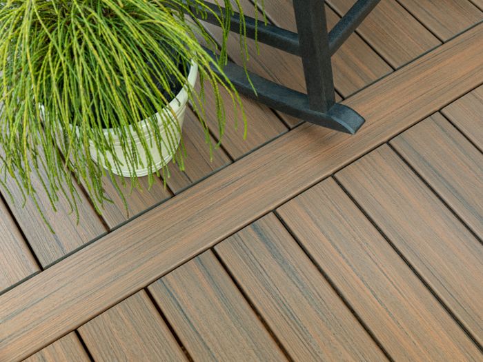 A detailed view of a wood-look composite deck features clean, parallel planks with a natural brown finish. A green potted plant sits on the deck, adding a touch of nature to the scene. The setting suggests an outdoor patio or backyard area, with a modern and tidy appearance. No visible text or numbers are present in the image.