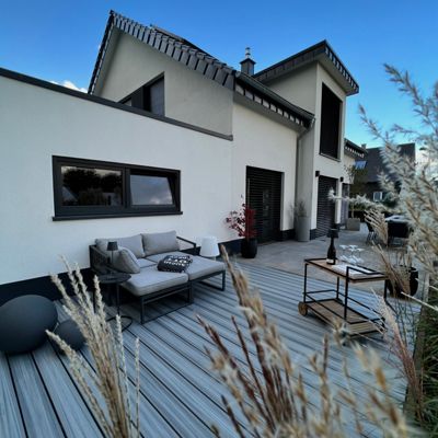 A contemporary house features a spacious outdoor patio with sleek gray furniture and a wooden deck. The setting includes a comfortable lounge area, a small table with a wine bottle and glasses, and decorative plants. The scene is captured in daylight with a clear blue sky, creating a relaxed and inviting atmosphere. No visible text or numbers are present in the image.