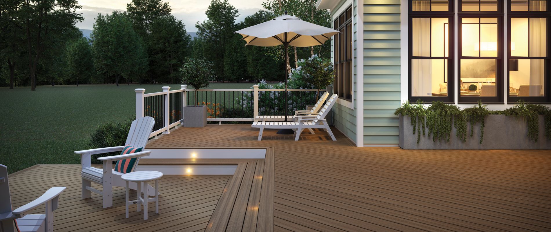 A spacious wooden deck extends from a house, featuring white Adirondack chairs and a table in the foreground. Lounge chairs and a large patio umbrella are set up near the railing, overlooking a lush green yard and trees. Warm lighting from the house windows creates a cozy atmosphere, while the deck is accented by built-in planters and soft evening light. The scene is tranquil, with no people present and a modern, inviting style.