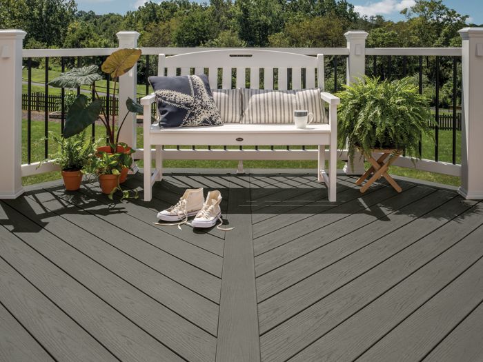 A modern outdoor deck features dark wood planks arranged in a chevron pattern. A white bench with striped cushions sits against a railing, surrounded by several potted plants and a pair of casual shoes. The setting is bright and inviting, with a clear blue sky and lush greenery in the background.