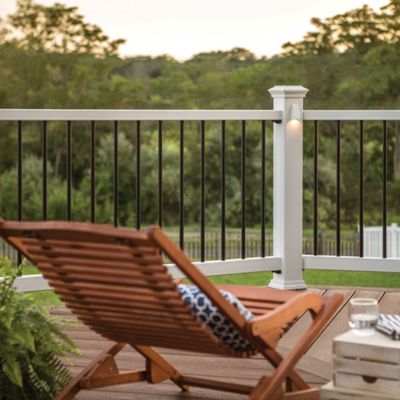 A wooden lounge chair with a patterned cushion is positioned on a wooden deck, facing a white and black railing. The setting is outdoors, overlooking lush greenery during sunset, creating a peaceful and inviting atmosphere. A small stack of books, a glass of water, and a potted fern add to the relaxed mood. Soft lighting on the railing posts enhances the cozy ambiance.