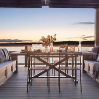 A stylish outdoor dining area is arranged on a wooden deck overlooking a tranquil lake at sunset. The scene features a table set with glassware, flowers, and a carafe, surrounded by cushioned benches with striped pillows. The soft evening light and pastel sky create a serene and inviting atmosphere, perfect for gatherings or relaxation.