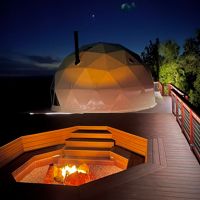 A modern geodesic dome structure is illuminated against a dark night sky, set on a wooden deck. In the foreground, a built-in fire pit with glowing flames creates a cozy atmosphere. The scene suggests a tranquil outdoor retreat, with warm lighting and natural surroundings visible.