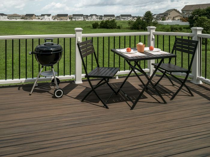 A modern outdoor deck features a small black charcoal grill and a compact patio dining set with two chairs and a table. The table holds two apples and a glass, set against a backdrop of green grass and suburban houses. White railings frame the deck, and the scene is bright with natural daylight, evoking a relaxed and inviting atmosphere.