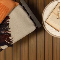 A marble-topped round side table holds an open book, a pair of eyeglasses, and a glass of amber liquid. The table sits beside a cushioned chair with a striped orange pillow and a dark fringed throw. The setting features warm wood flooring, creating a relaxed and inviting atmosphere.